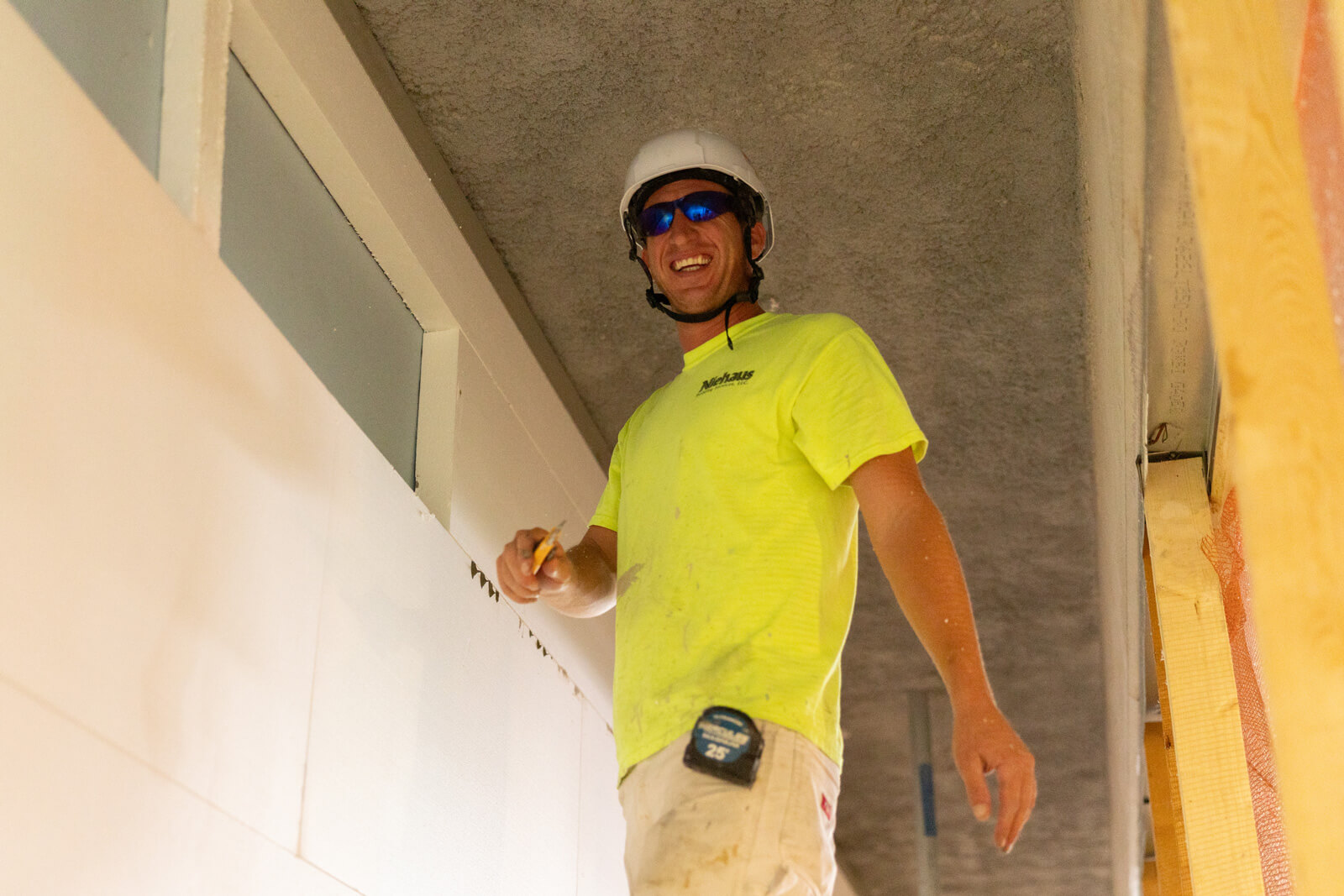 Construction worker in safety glasses and hard hat measuring interior wall panels near a fireproofed ceiling.