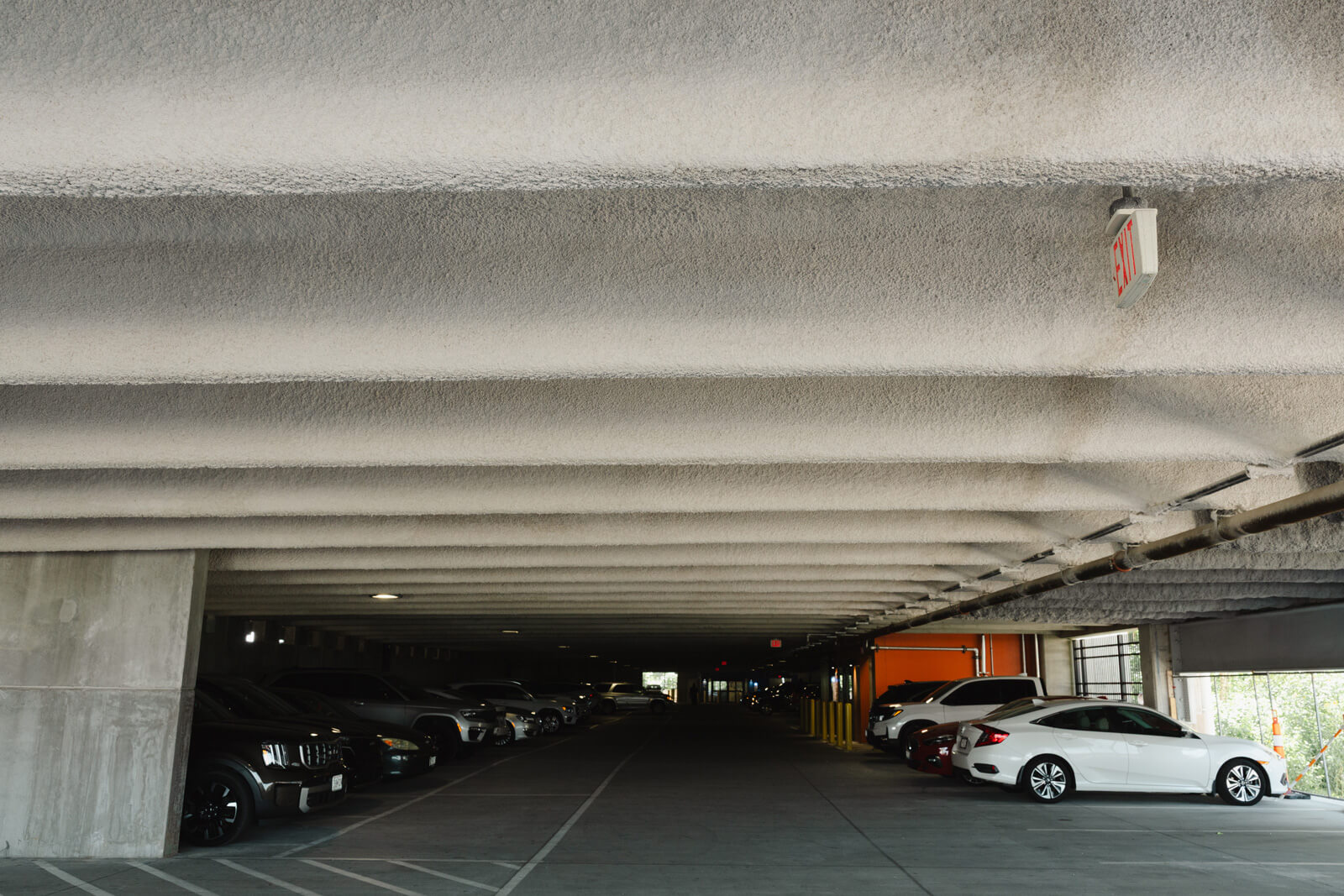 Covered parking garage displaying uniform spray-applied fireproofing on concrete ceiling beams above parked vehicles.